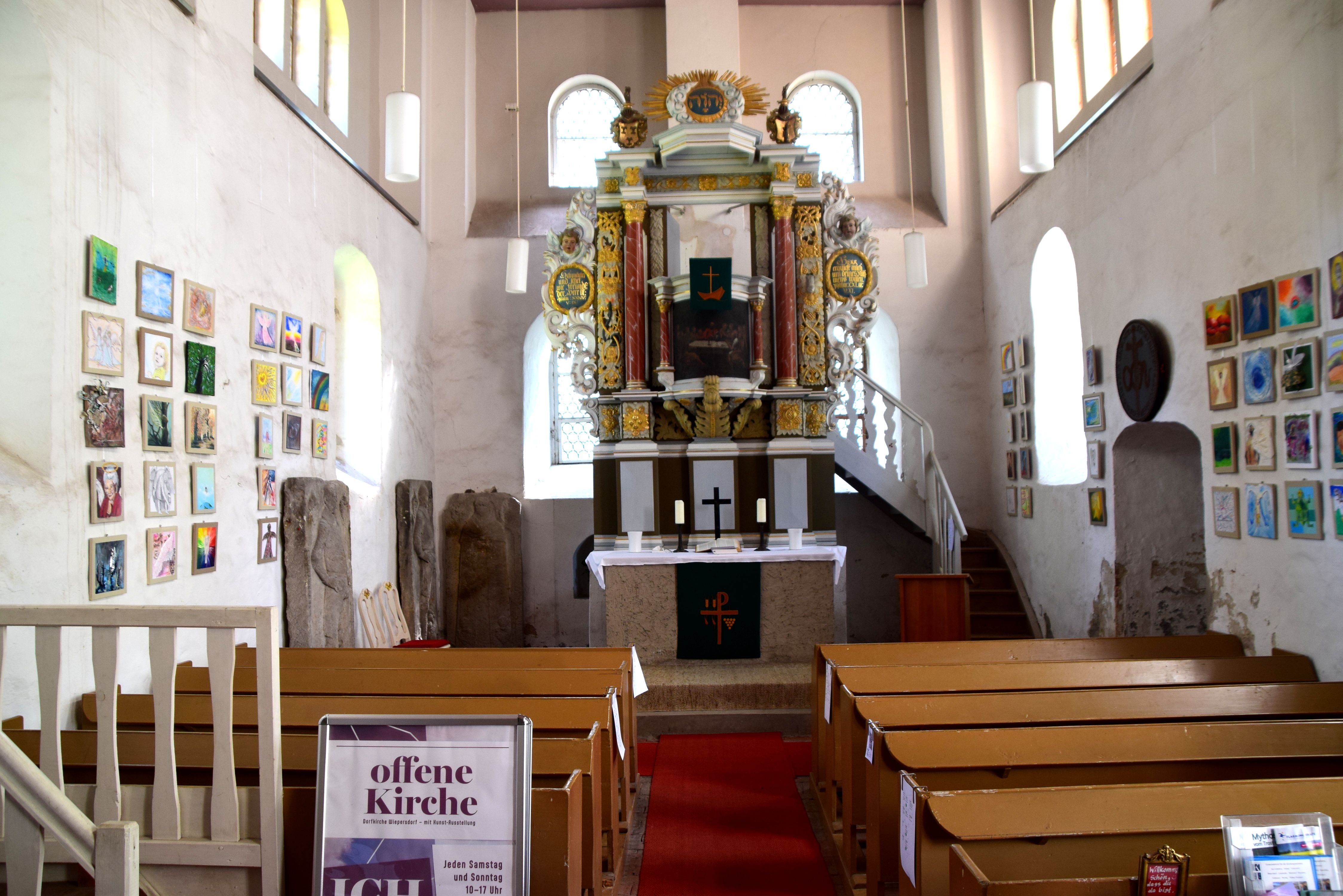 Innenraum der Kirche in Wiepersdorf. An den Wänden hängen Grafiken und Gemälde der Enges-Aisstellung von 2023-24. Die Werke wurden inzwischen für einen guten Zweck versteigert.